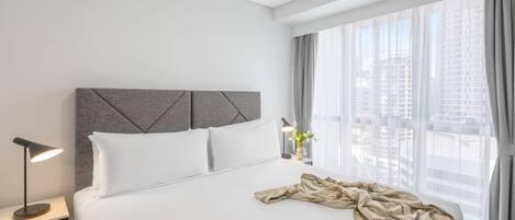 A hotel room with a neatly made bed, a bedside lamp, and a large window with curtains.