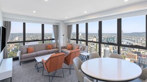 Une chambre d’hôtel moderne dotée d’une grande fenêtre donnant sur la ville, d’un ensemble de canapés, d’une table à manger et d’un téléviseur à écran plat.