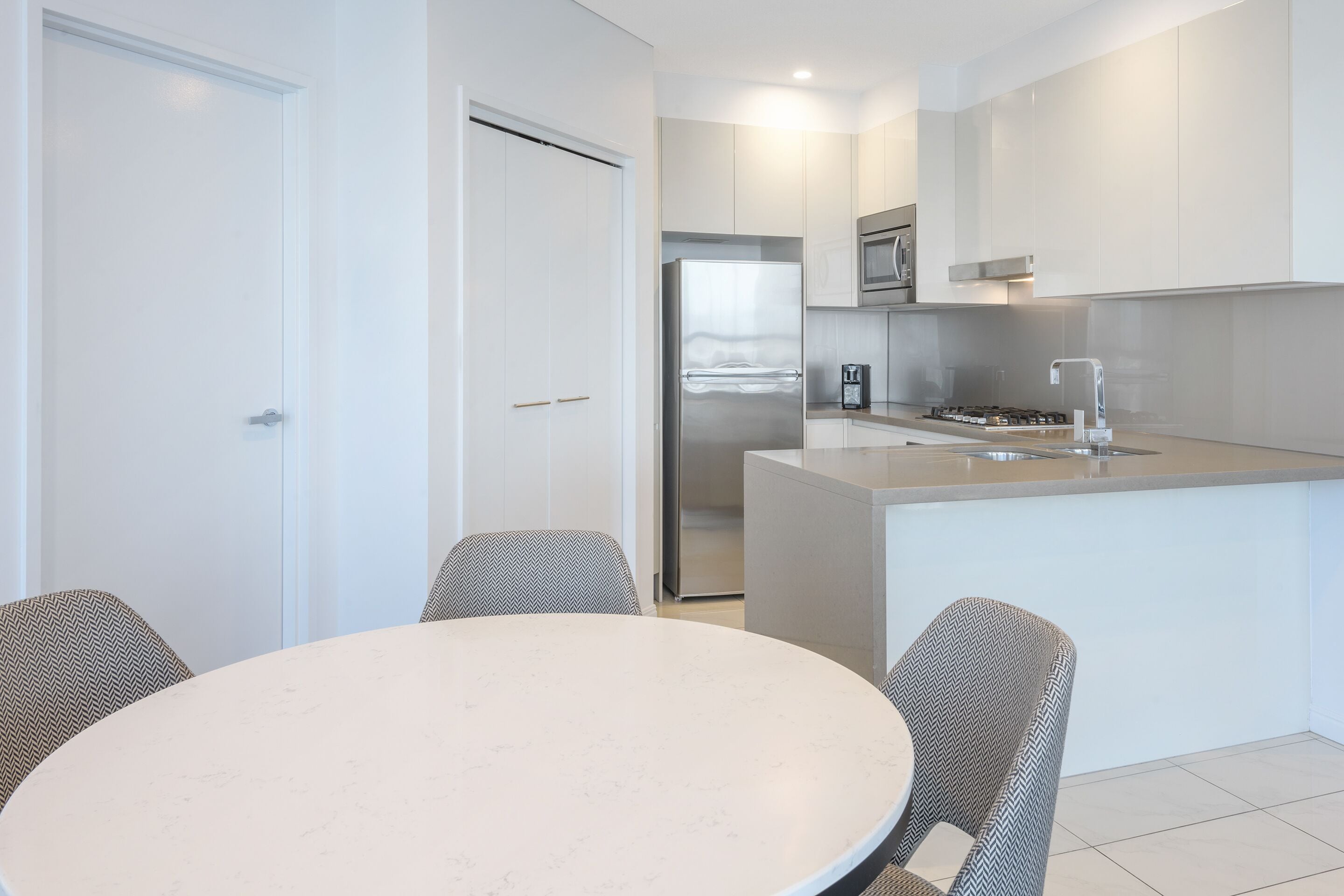 Een moderne keuken met witte kasten, een roestvrijstalen koelkast en een inbouwoven. Er is een eethoek met een ronde tafel en twee stoelen.