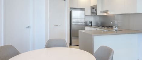 A modern kitchen with white cabinetry, a stainless steel refrigerator, and a built-in oven. There is a dining area with a round table and two chairs.