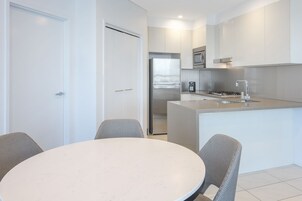 A modern kitchen with white cabinetry, a stainless steel refrigerator, and a built-in oven. There is a dining area with a round table and two chairs.