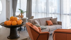 Un salon moderne avec un canapé, une table à manger agrémentée d’un panier de fruits, et une vue sur le paysage urbain à travers de grandes fenêtres.