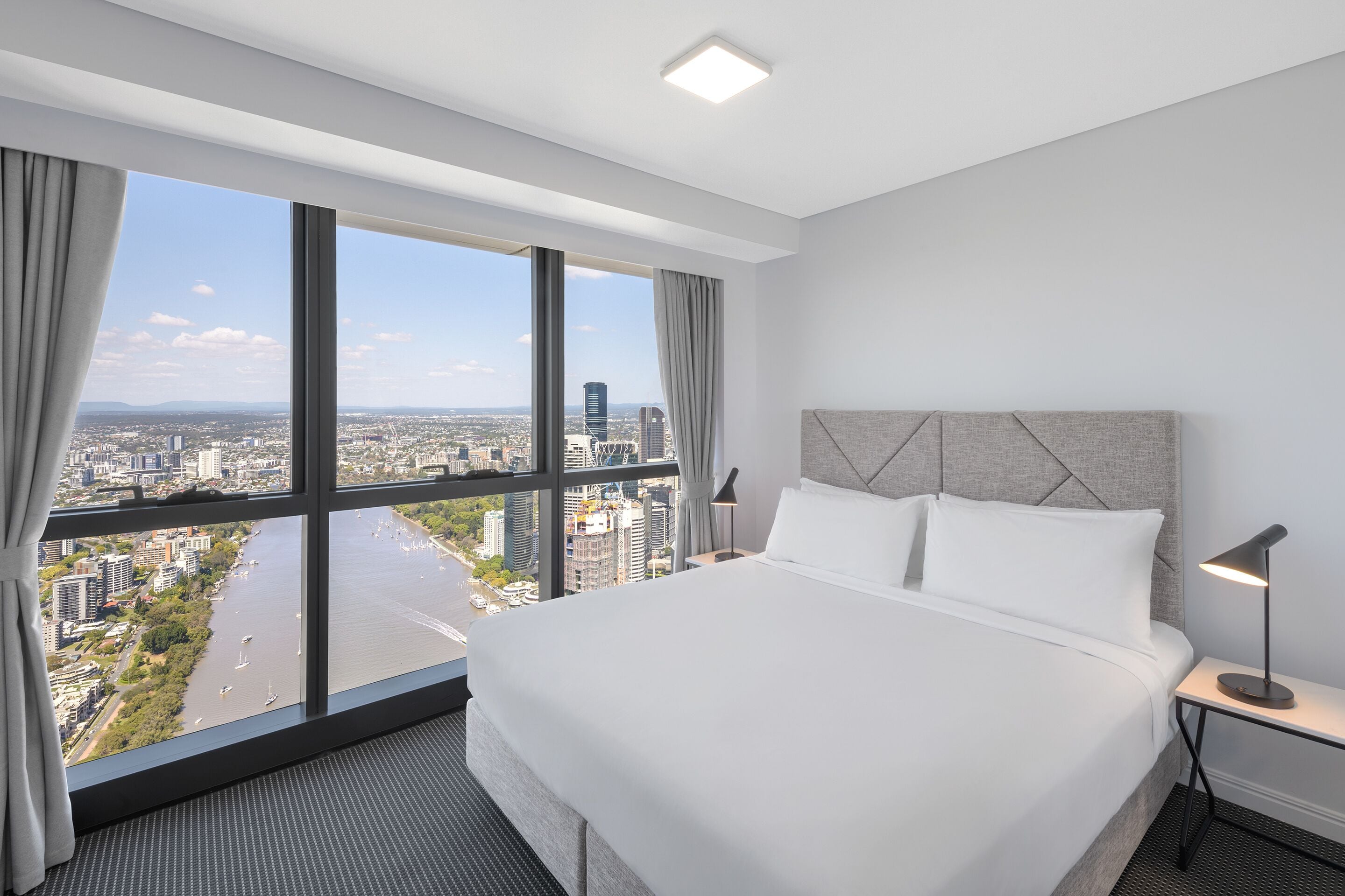 A hotel room with a large bed, a nightstand, a lamp, and a view of a cityscape and river.