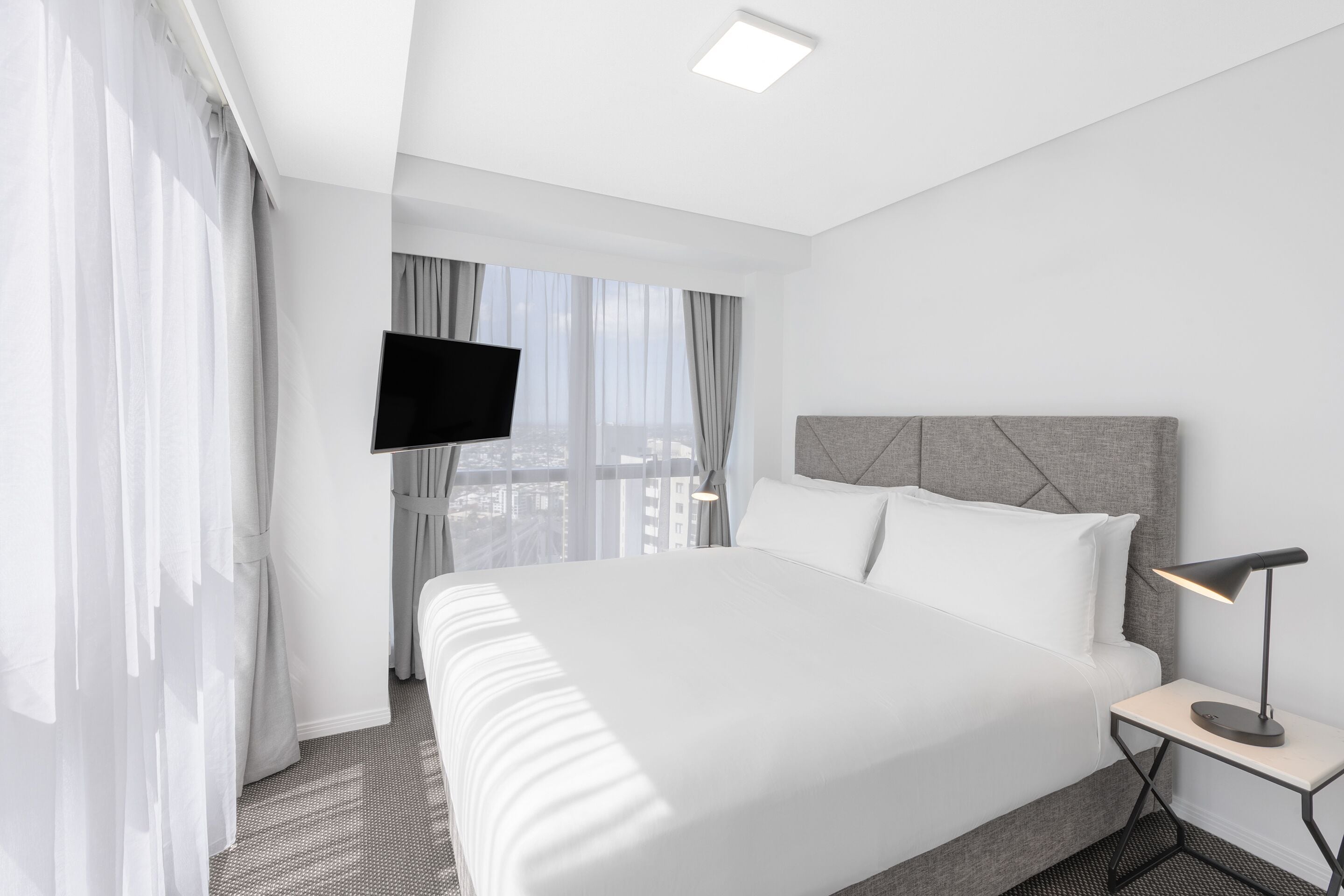 A modern hotel room with a large bed, a flat-screen TV mounted on the wall, and a bedside table with a lamp.