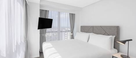 A modern hotel room with a large bed, a flat-screen TV mounted on the wall, and a bedside table with a lamp.