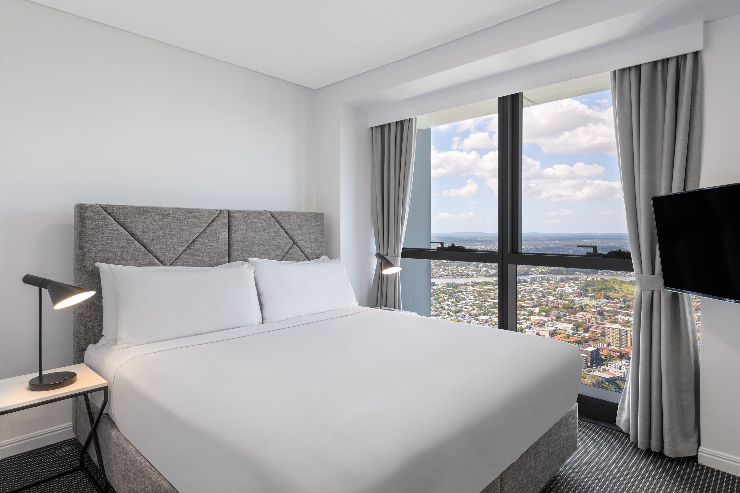 Une chambre d’hôtel avec un grand lit, une table de chevet avec une lampe, une télévision et une fenêtre donnant sur la ville.