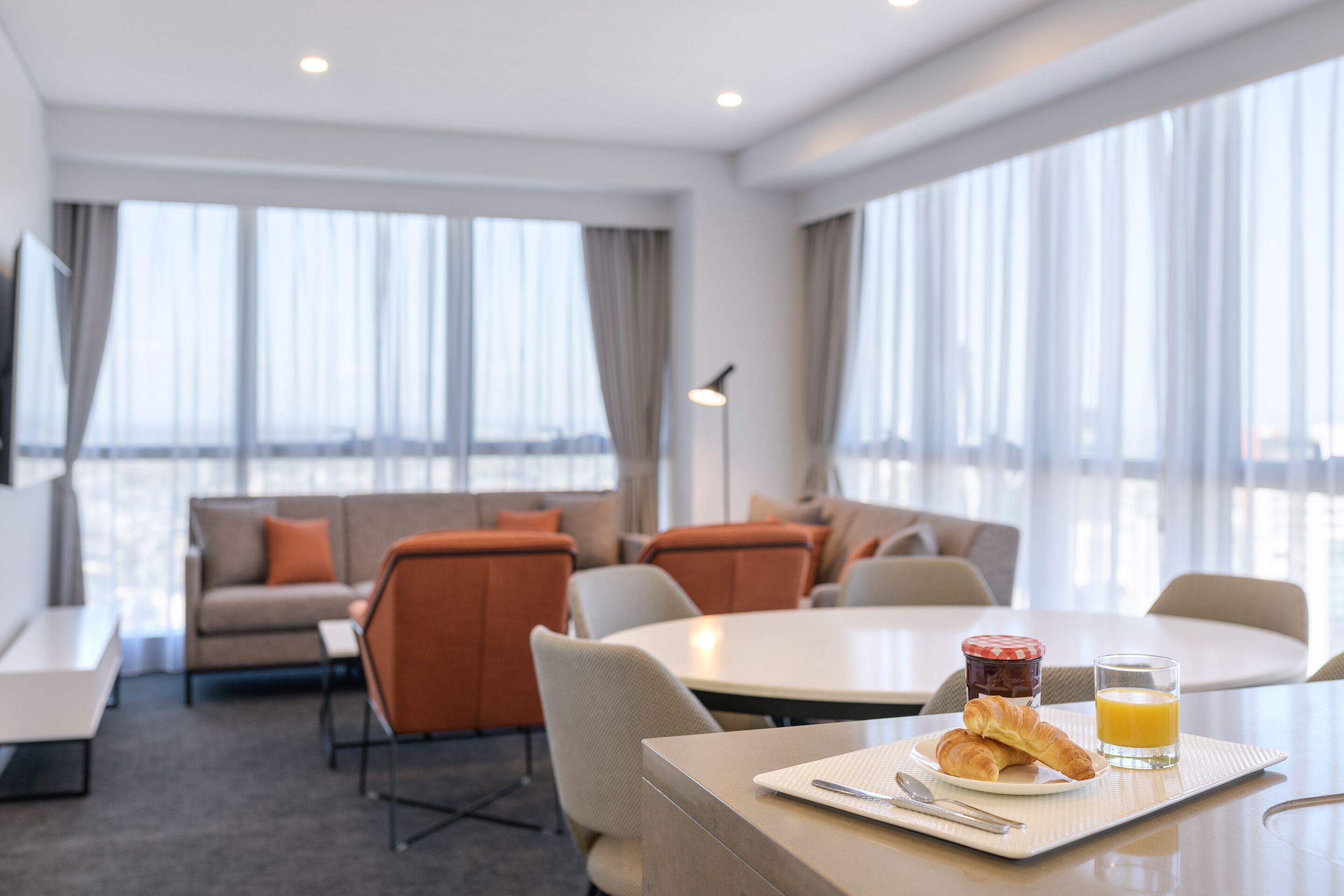 Una habitación de hotel moderna con comedor, un conjunto de sofás y una mesa para el desayuno con croissants y jugo de naranja.