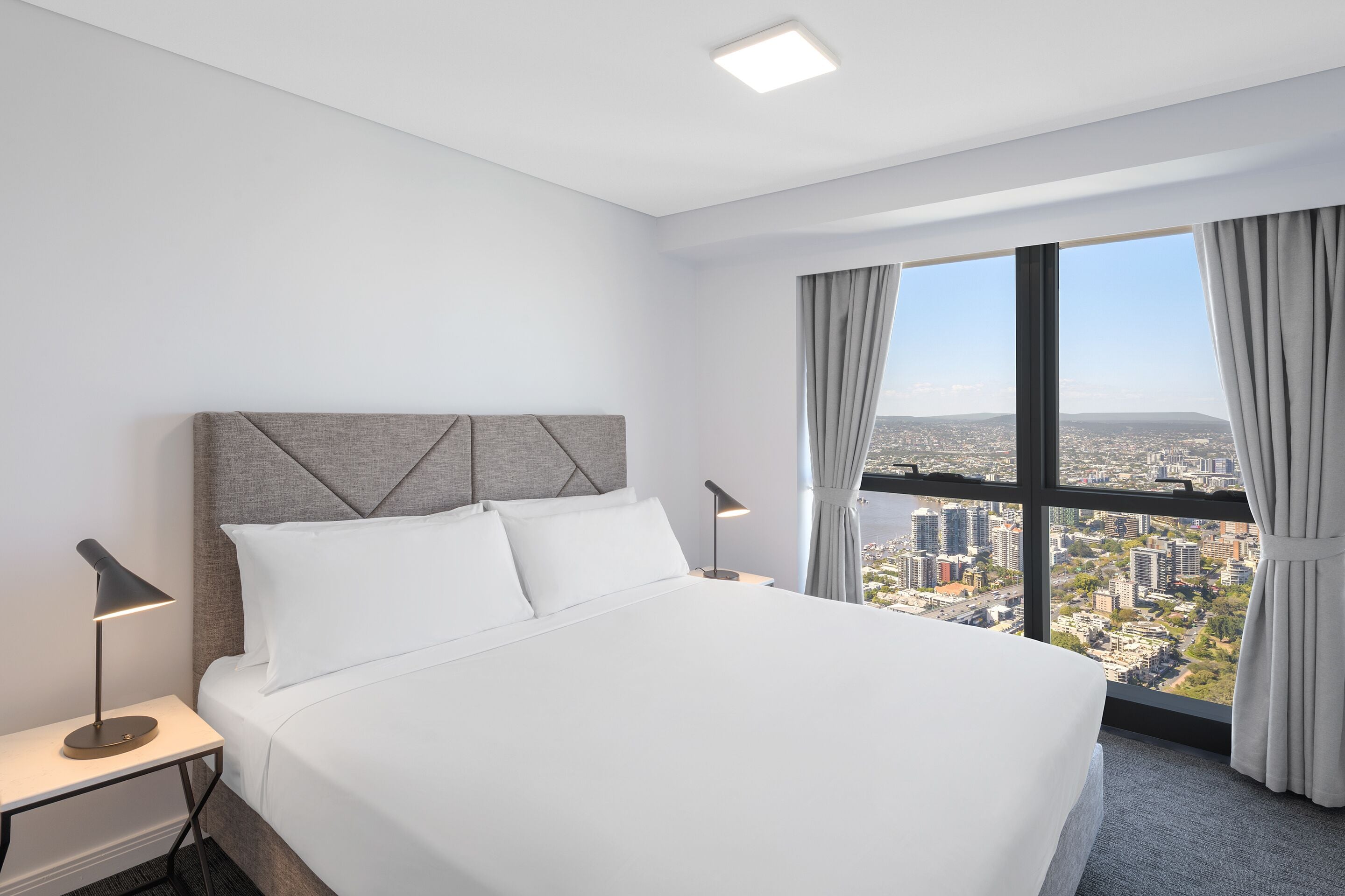 A hotel room with a large bed, two bedside tables with lamps, a view of a cityscape through large windows, and a modern ceiling light.