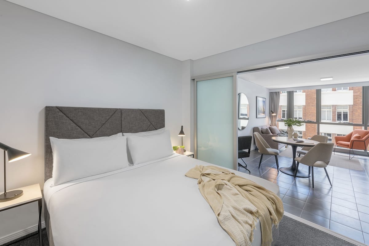 Habitación de hotel moderna con una cama grande, una mesita de noche, una lámpara y vistas de la ciudad a través de amplias ventanas.