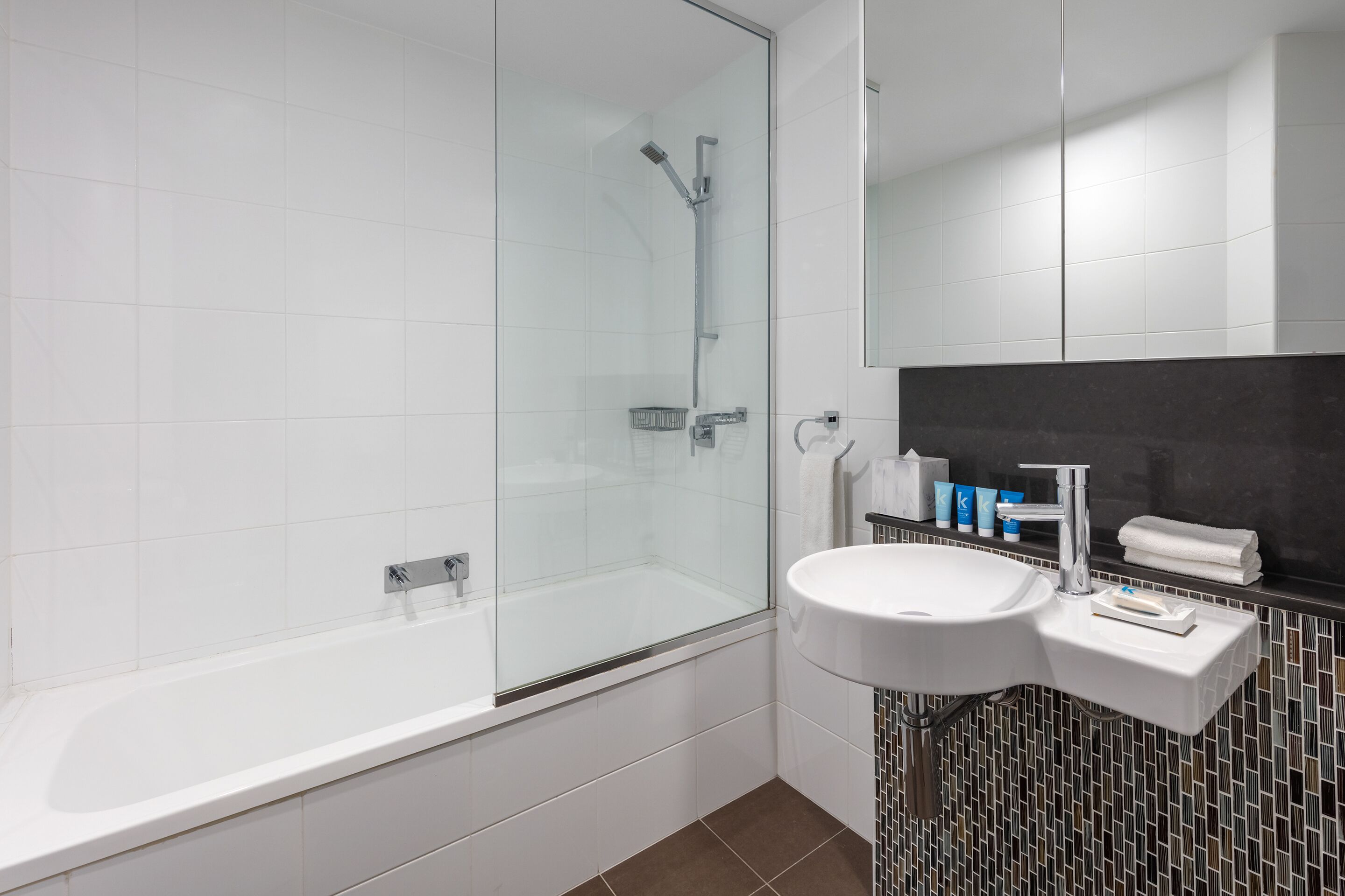 A modern bathroom with a glass-enclosed shower, a white sink with a black tiled backsplash, and a mirror above the sink.
