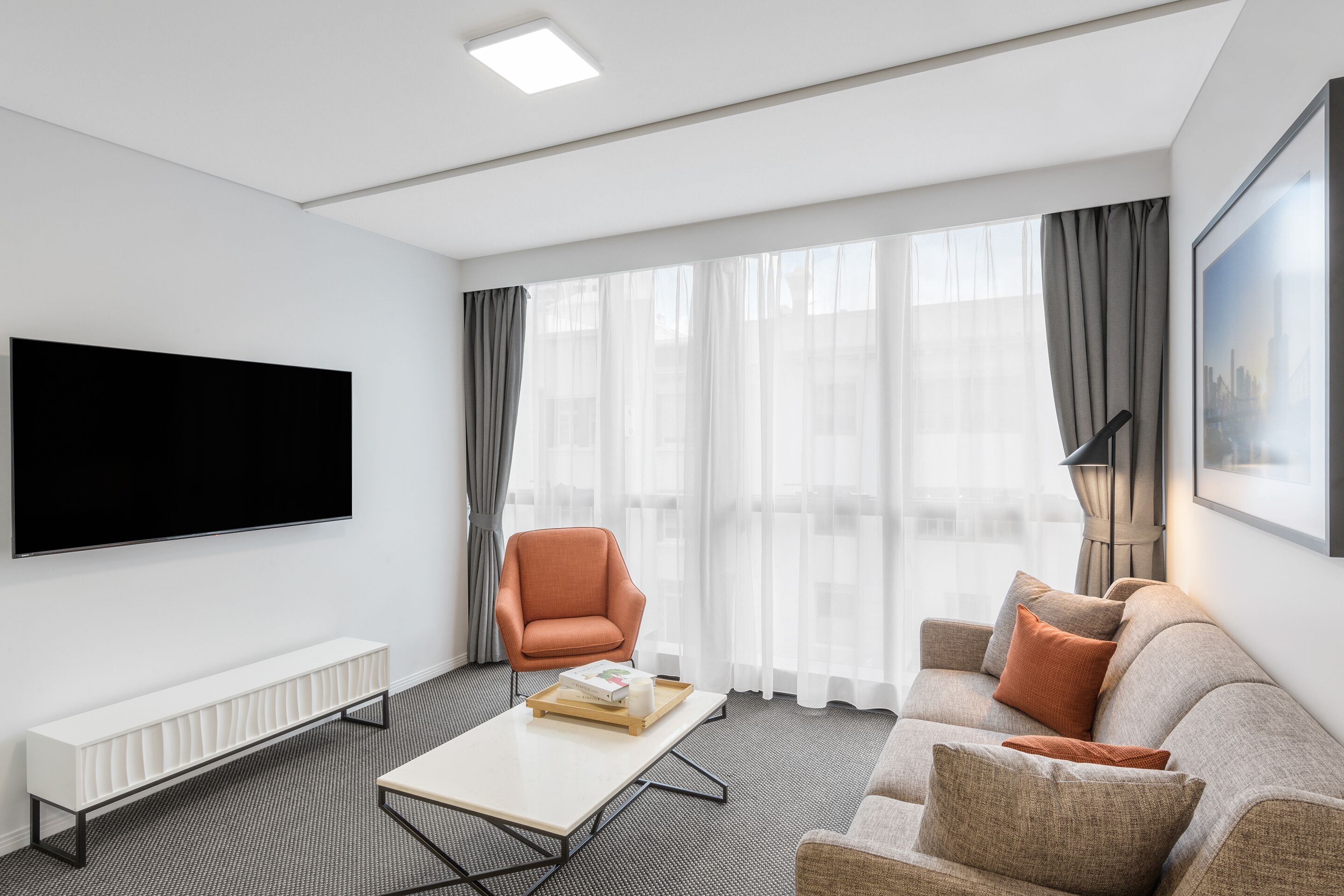 A modern living room with a flat-screen TV, a white console table, a grey sofa with orange cushions, and a grey armchair.