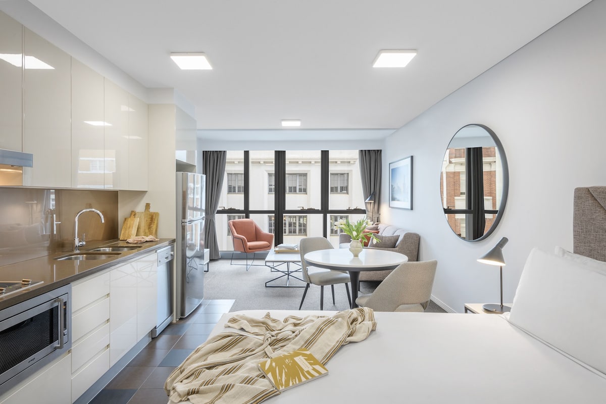 Habitación de hotel moderna con kitchenette, zona de comedor, una cama grande y vistas a la ciudad a través de amplios ventanales.