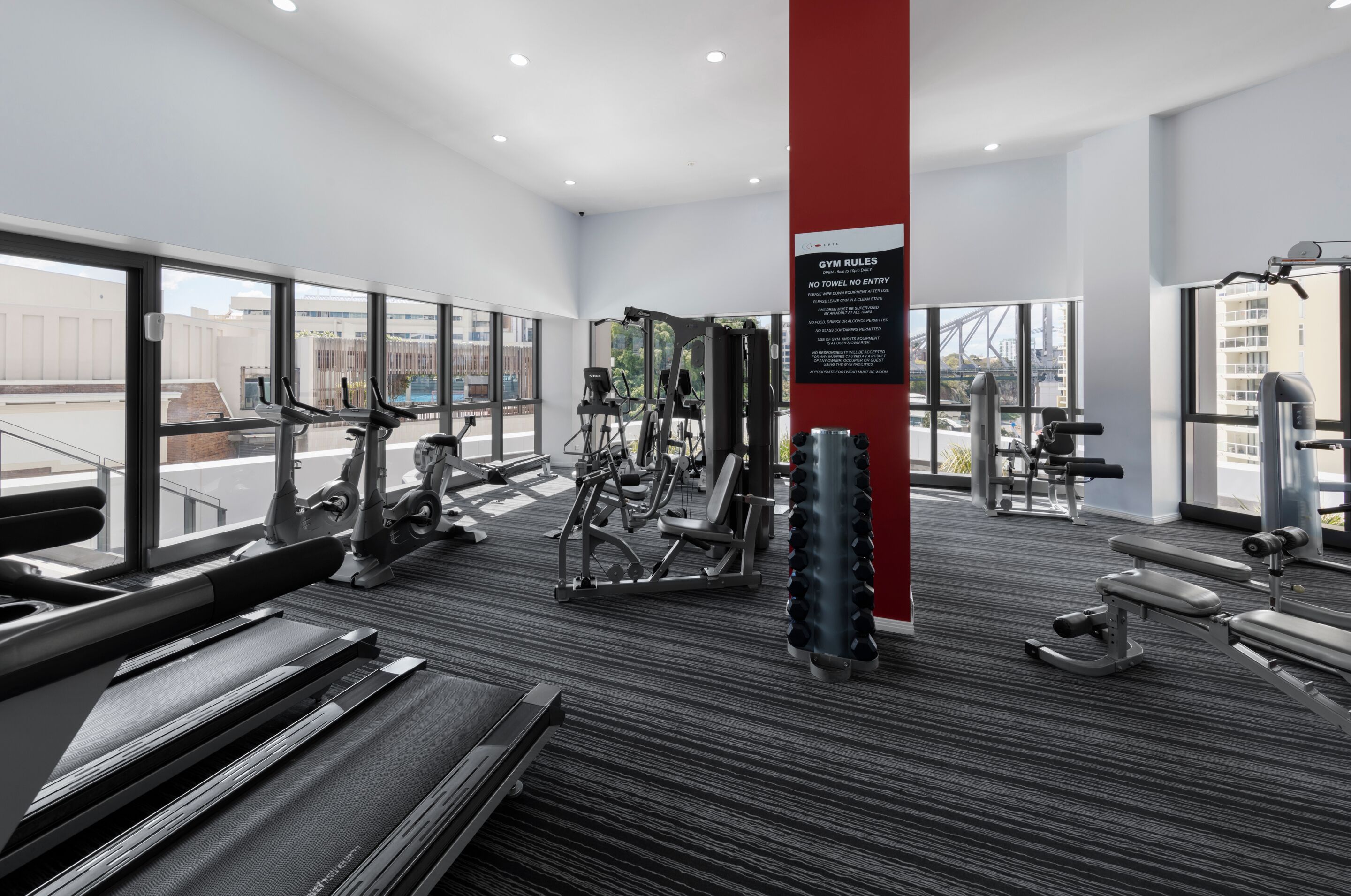 A modern gym with cardio machines, free weights, and a red pillar with gym rules.