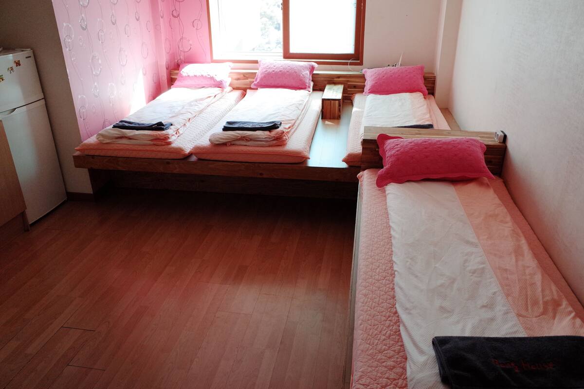 Une chambre avec deux lits en bois, chacun agrémenté de draps roses et blancs. Une petite table en bois se trouve entre les lits, et un réfrigérateur blanc est placé à gauche.