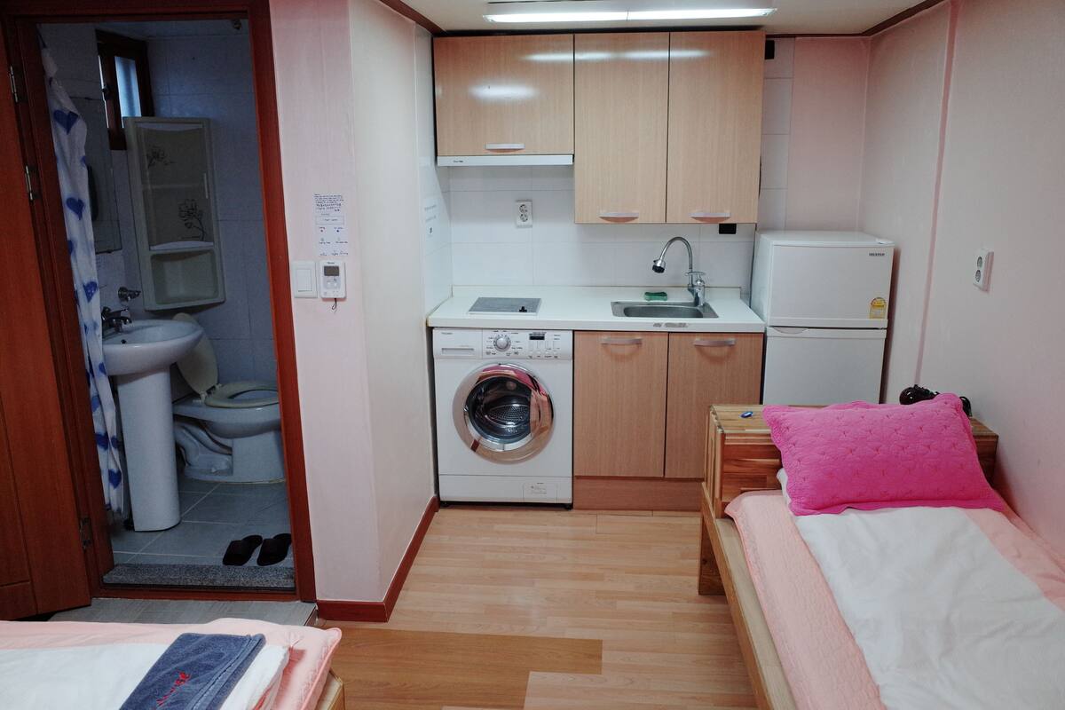 Une petite pièce comprenant un lit, un lave-linge, un évier et un petit réfrigérateur.