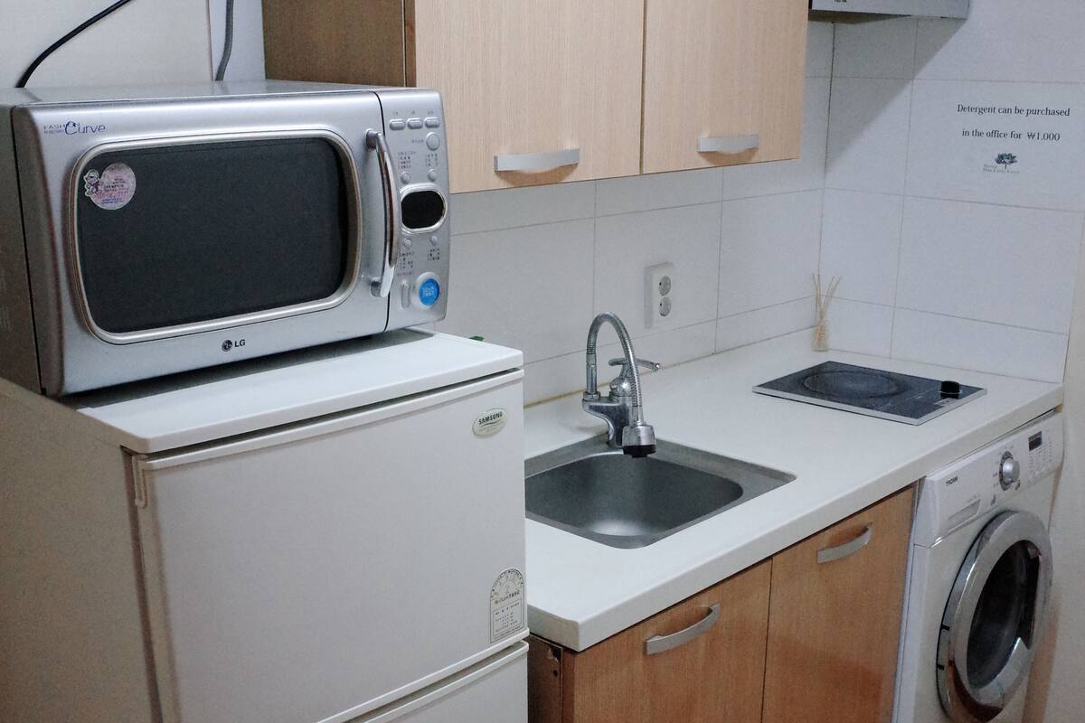 Une cuisine compacte avec un micro-ondes posé sur le dessus d’un réfrigérateur, un évier, un lave-linge et des placards en bois.