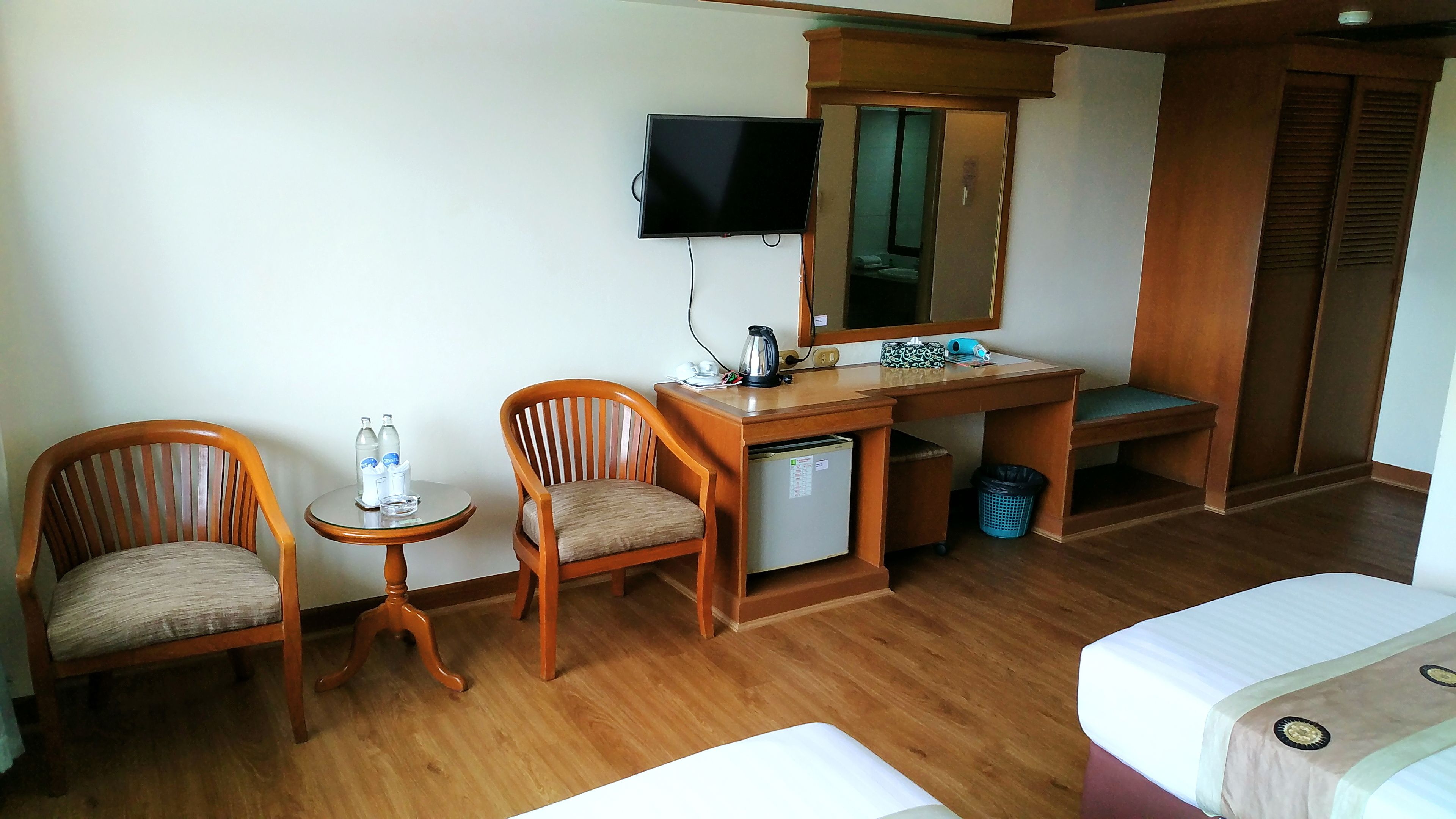 Habitación de hotel con dos sillas de madera, una mesita con botellas de agua, un escritorio con espejo, un televisor y una cama.