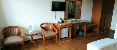 Habitación de hotel con dos sillas de madera, una mesita con botellas de agua, un escritorio con espejo, un televisor y una cama.