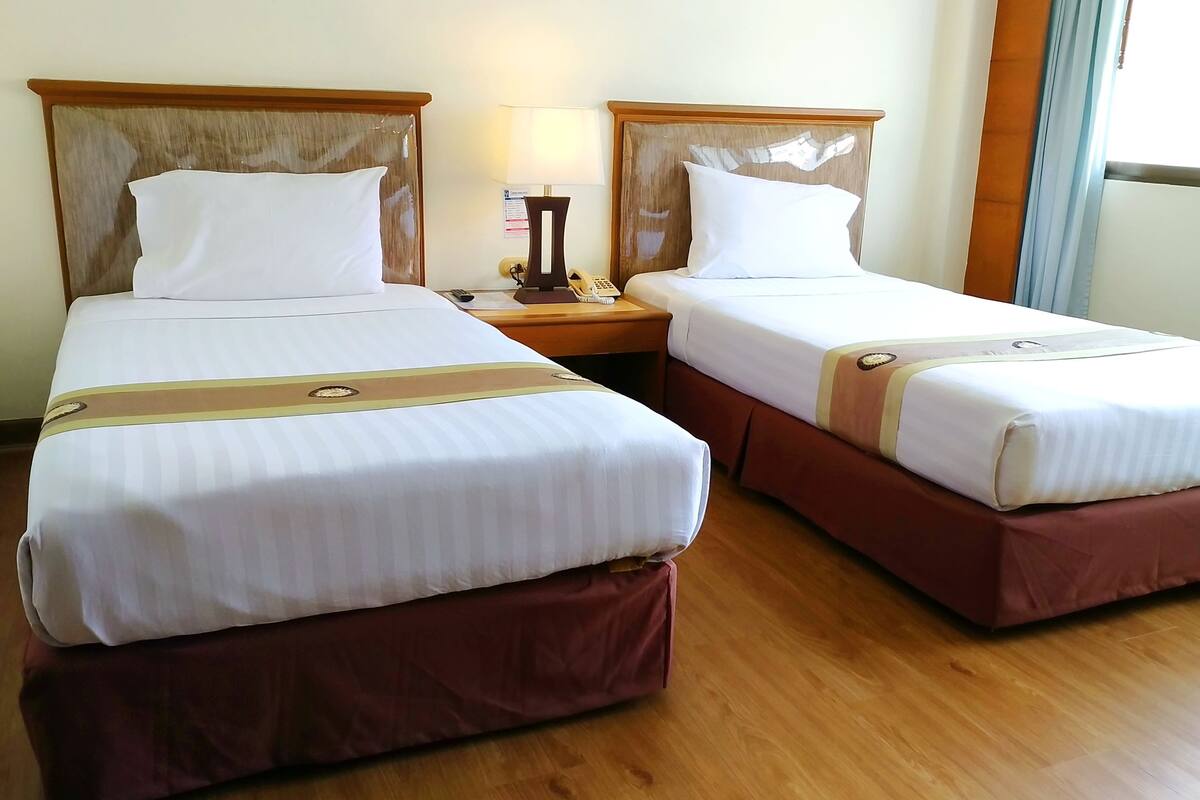 Two beds with wooden headboards, a bedside table with a lamp, and a window with curtains.