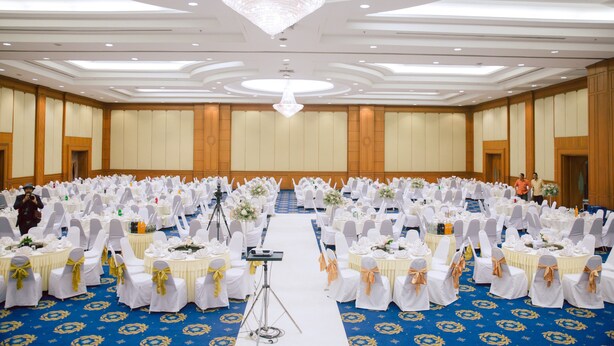 A large banquet hall with rows of round tables, white tablecloths, and gold sashes. - Grand Park Hotel (Nakhon Si Thammarat)