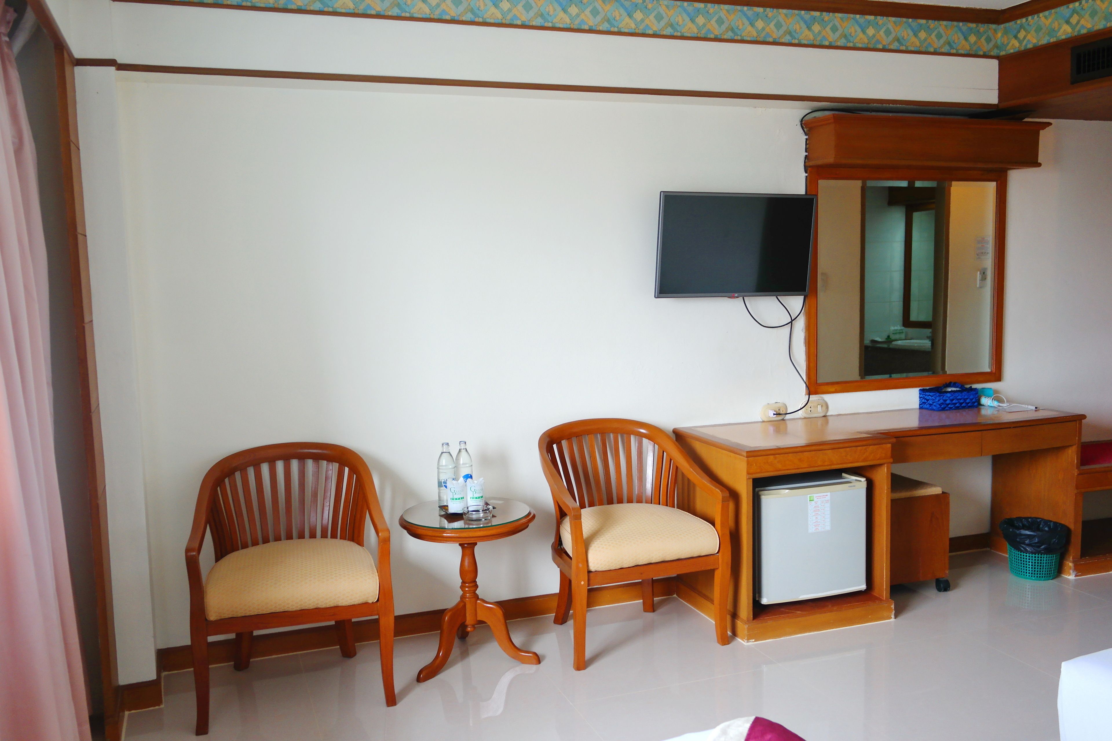 A hotel room with two wooden chairs, a small round table with water bottles, a television mounted on the wall, and a small refrigerator under the desk.
