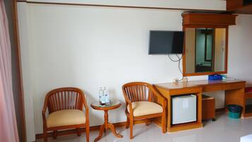 A hotel room with two wooden chairs, a small round table with water bottles, a television mounted on the wall, and a small refrigerator under the desk.