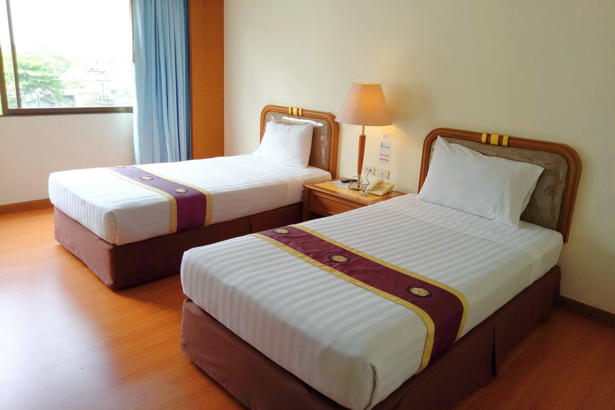 A hotel room with two single beds, wooden headboards, a bedside table with a lamp, and a window with curtains.