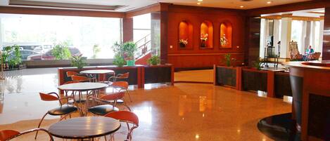 A spacious lobby with round tables and orange chairs, a reception desk, and a view of parked cars outside.