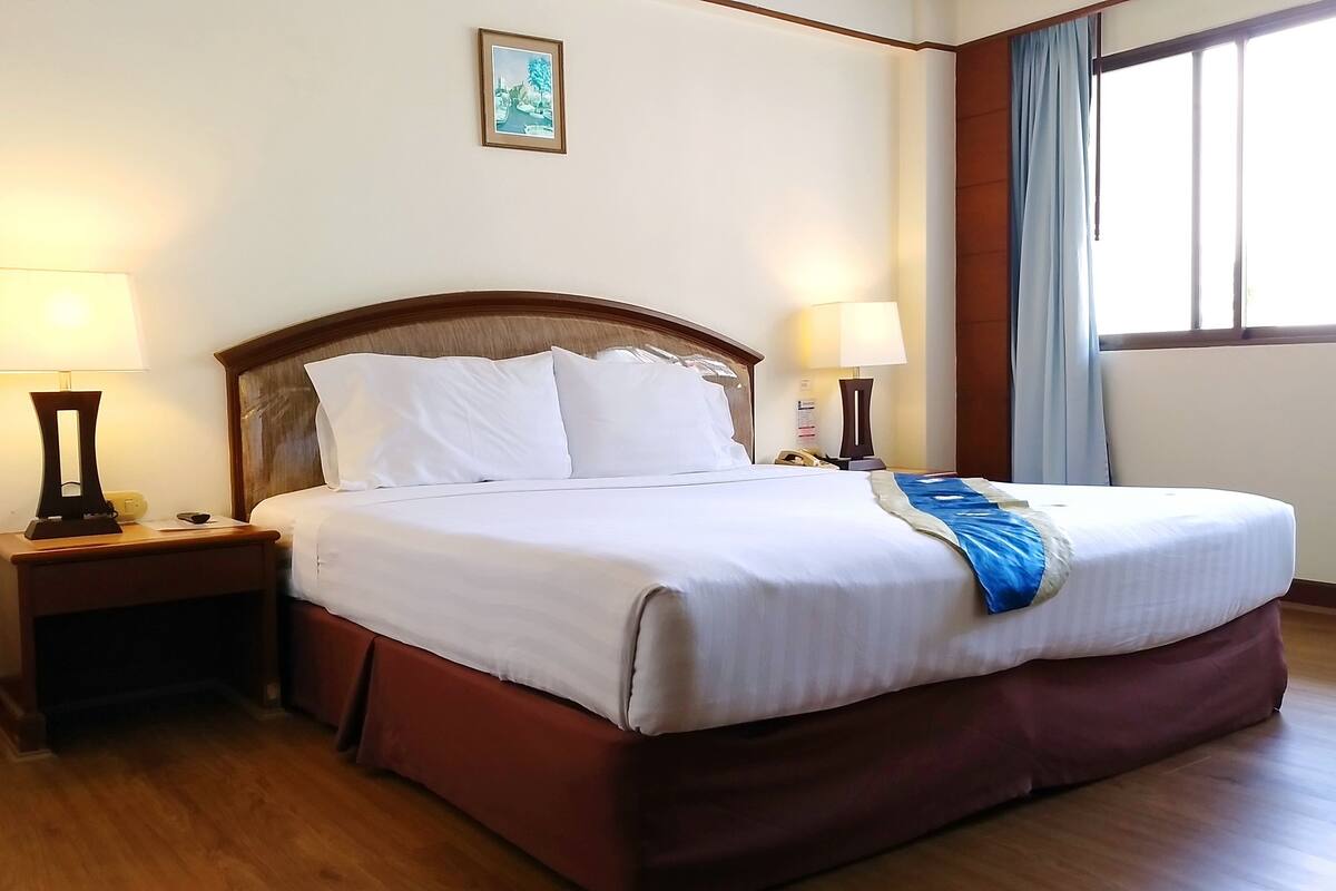 A neatly made bed with white linens, two bedside lamps, a wooden headboard, and a window with curtains.