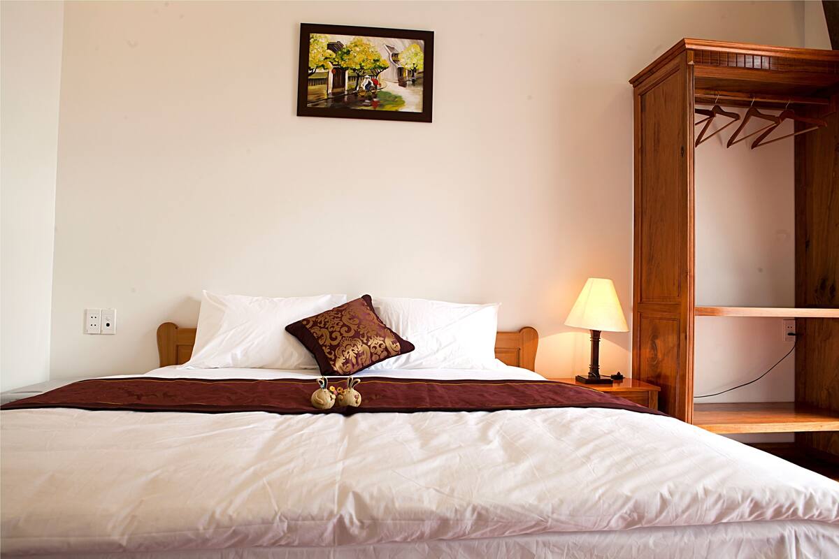Ein ordentlich bezogenes Bett mit weißen Laken, einer braunen Tagesdecke und einem Zierkissen. Ein Holznachttisch mit Lampe und Steckdose ist vorhanden. Rechts ist ein Holzschrank mit Kleiderstange zu sehen.