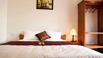Cama bem arrumada com lençóis brancos, manta marrom e almofada decorativa. Um criado-mudo de madeira com luminária e tomada. Guarda-roupa de madeira com cabideiro visível à direita.