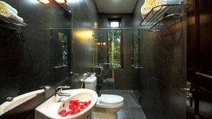Un bany modern amb dutxa de vidre, un lavabo blanc amb pètals de rosa, un vàter i parets de rajola fosca.