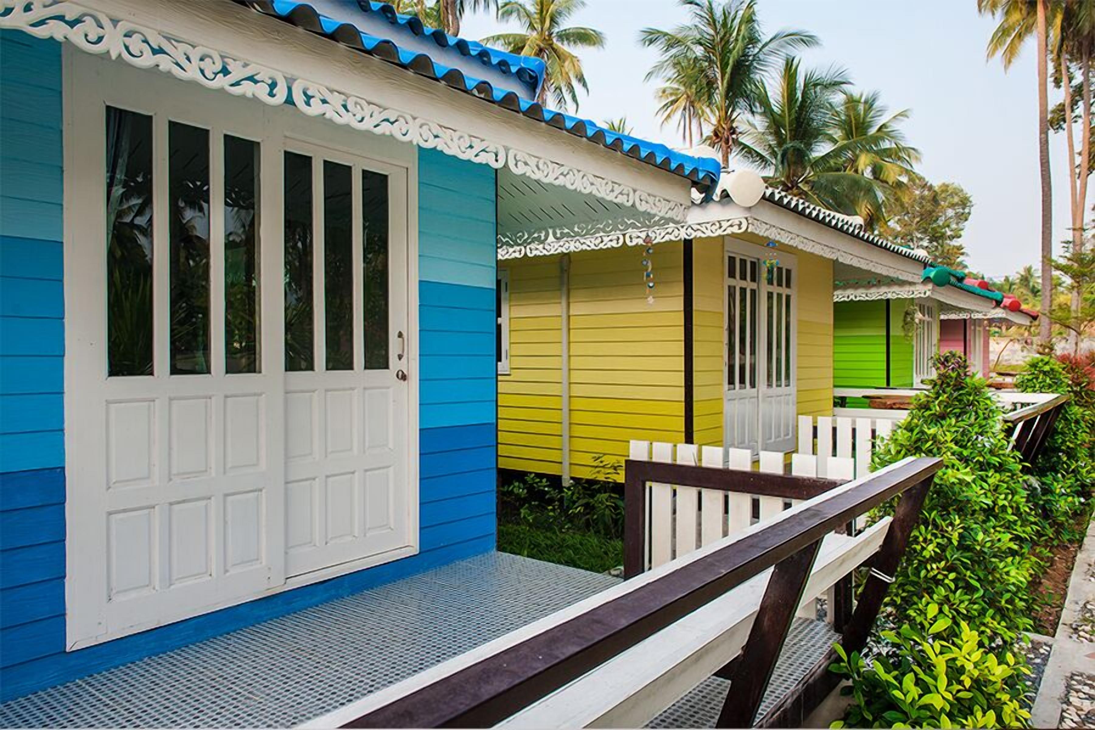 Una hilera de cabañas de un piso, coloridas, con ventanas y puertas enmarcadas en blanco, un porche con baranda de madera y un fondo de palmeras.