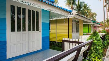 Una hilera de cabañas de un piso, coloridas, con ventanas y puertas enmarcadas en blanco, un porche con baranda de madera y un fondo de palmeras.