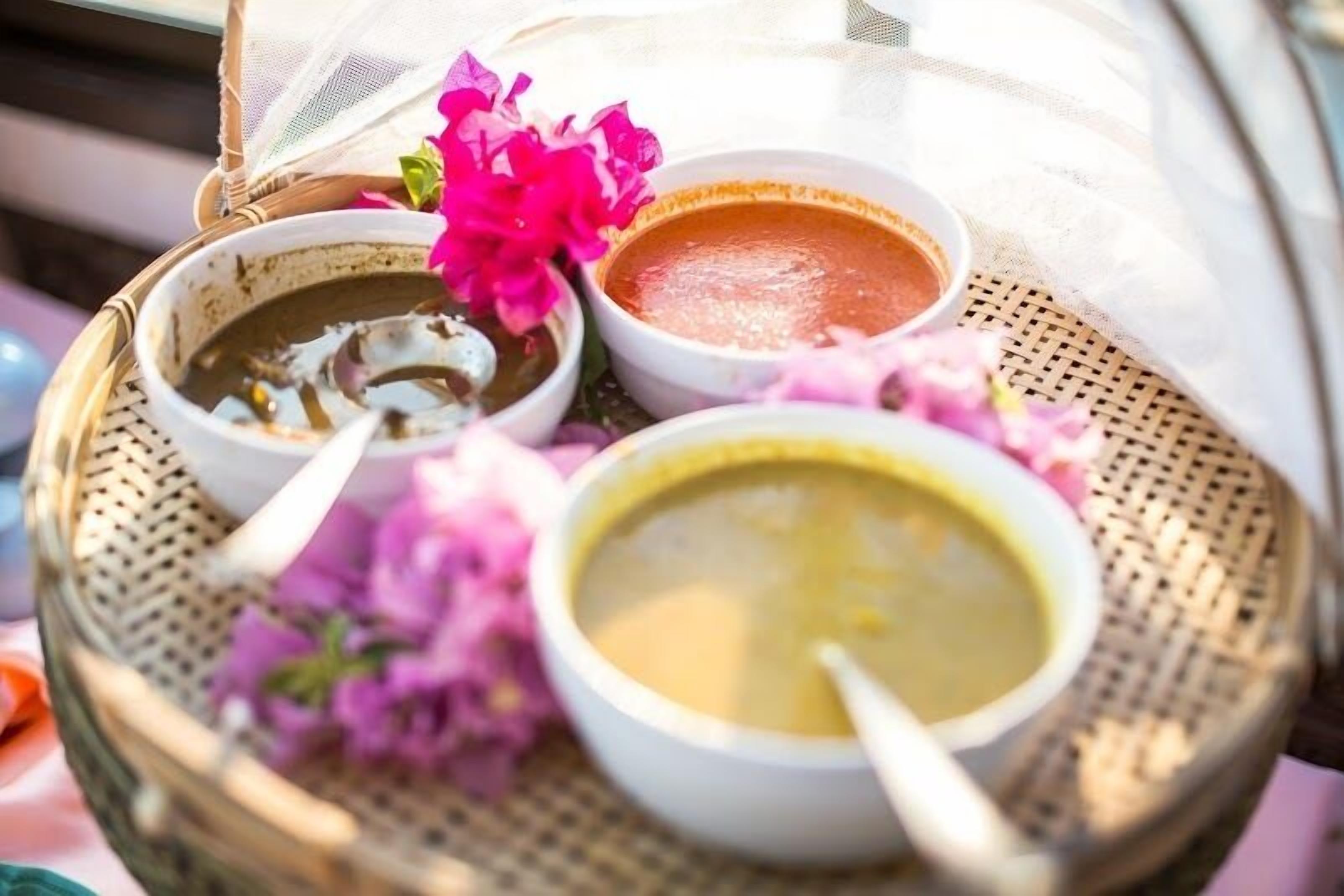 Una bandeja con tres tazones de sopa, cada uno con una cuchara, y una cesta con flores.