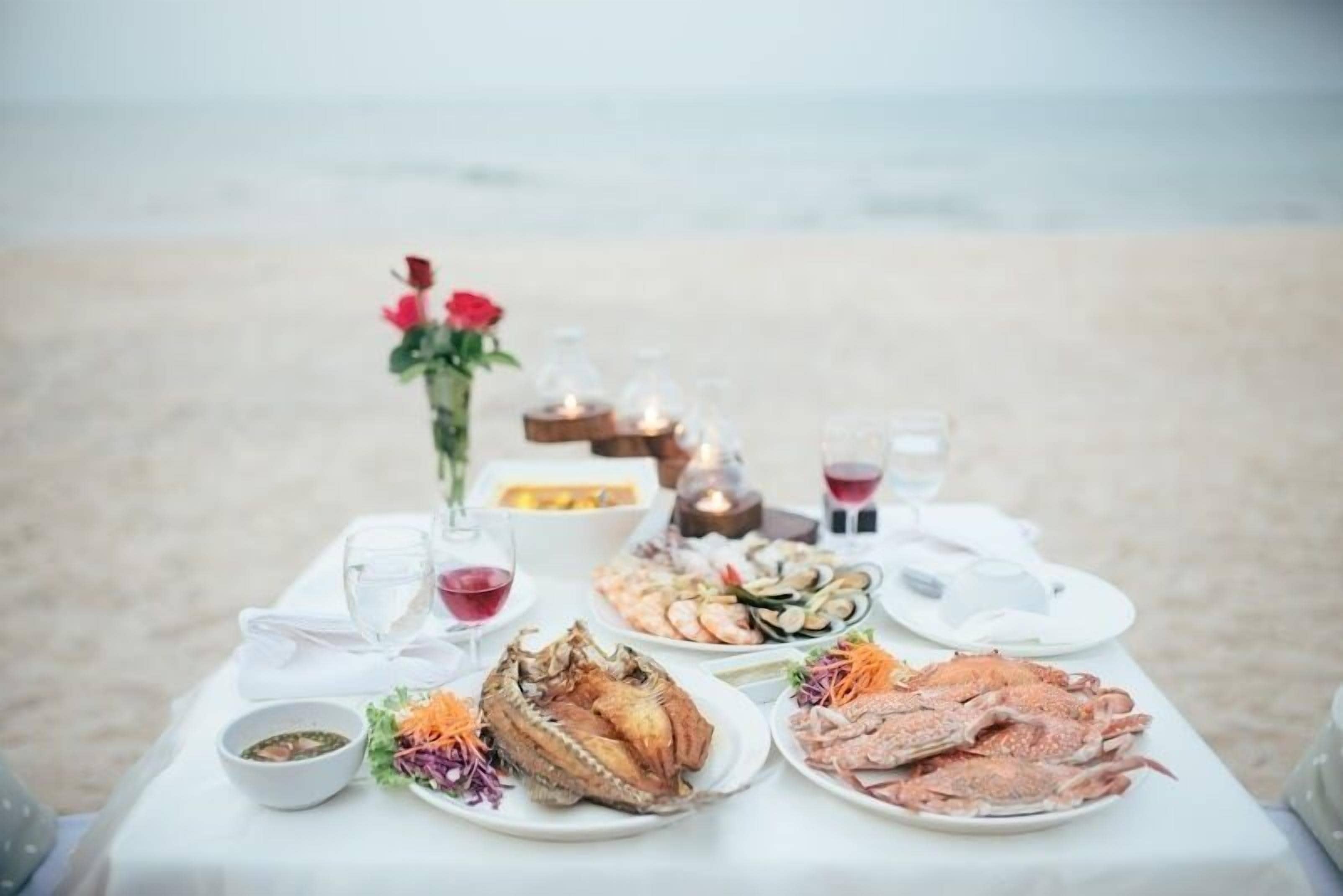 A table set on a beach with seafood dishes, glasses, and a vase with flowers.