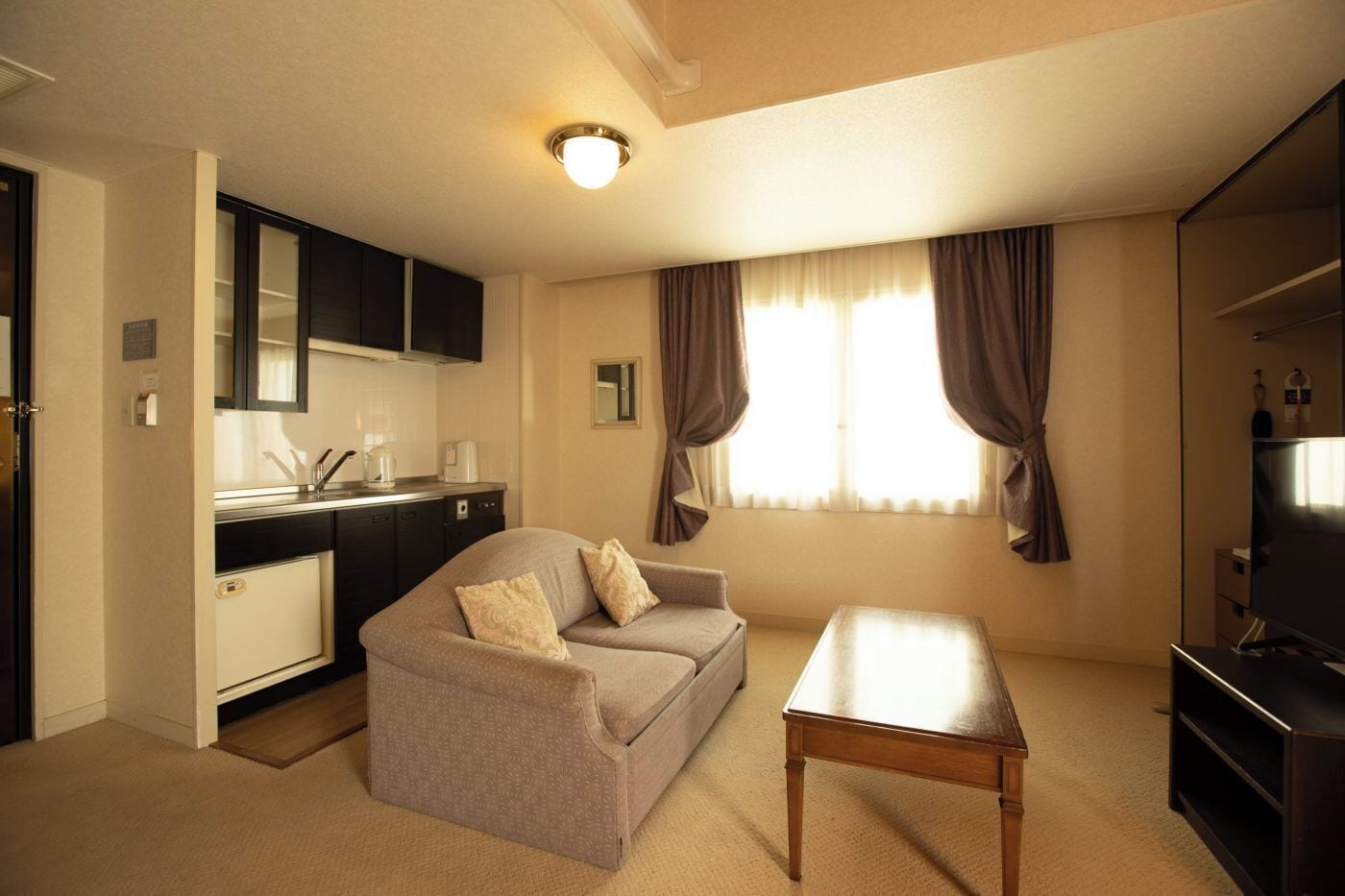 Un espacio habitable compacto con sofá, una mesa de centro, televisor, kitchenette y una ventana con cortinas.