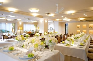 Une salle de banquet avec de longues tables dressées pour un événement formel, agrémentée de nappes blanches, de compositions florales et d’un lustre.