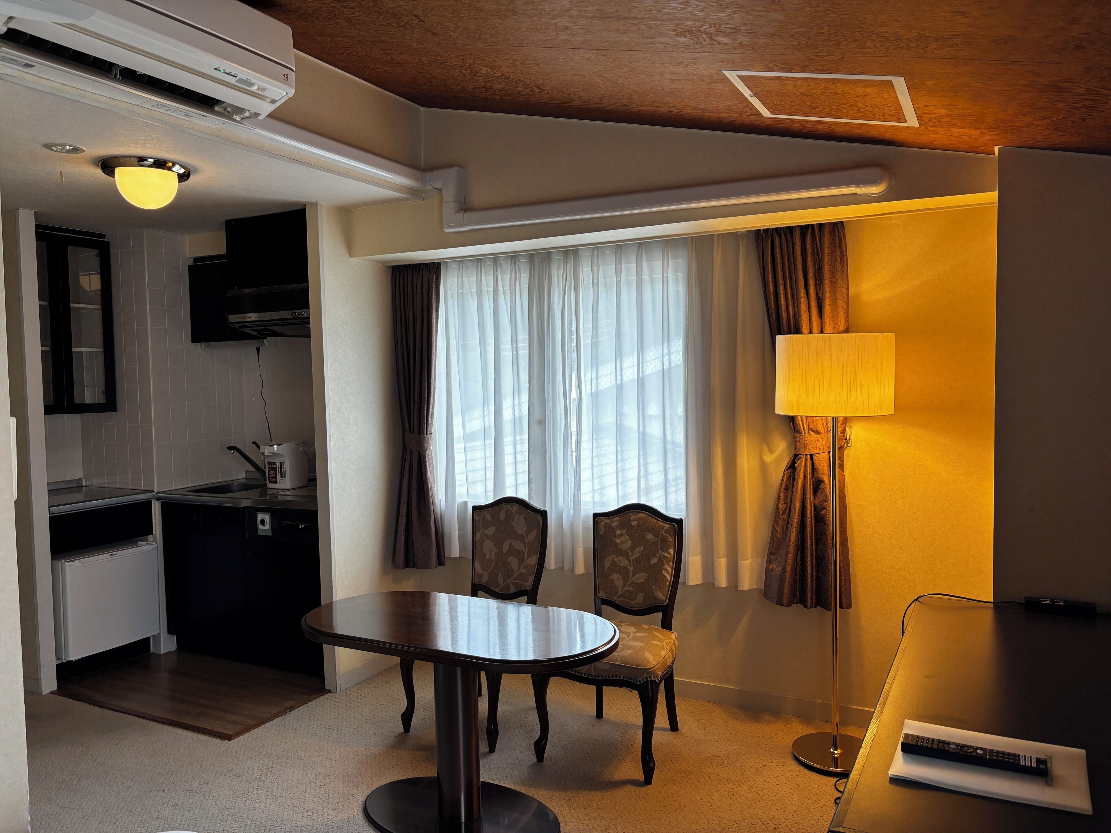 Habitación de hotel con una mesa de comedor, dos sillas, una lámpara, un televisor y una kitchenette con microondas y hervidor.