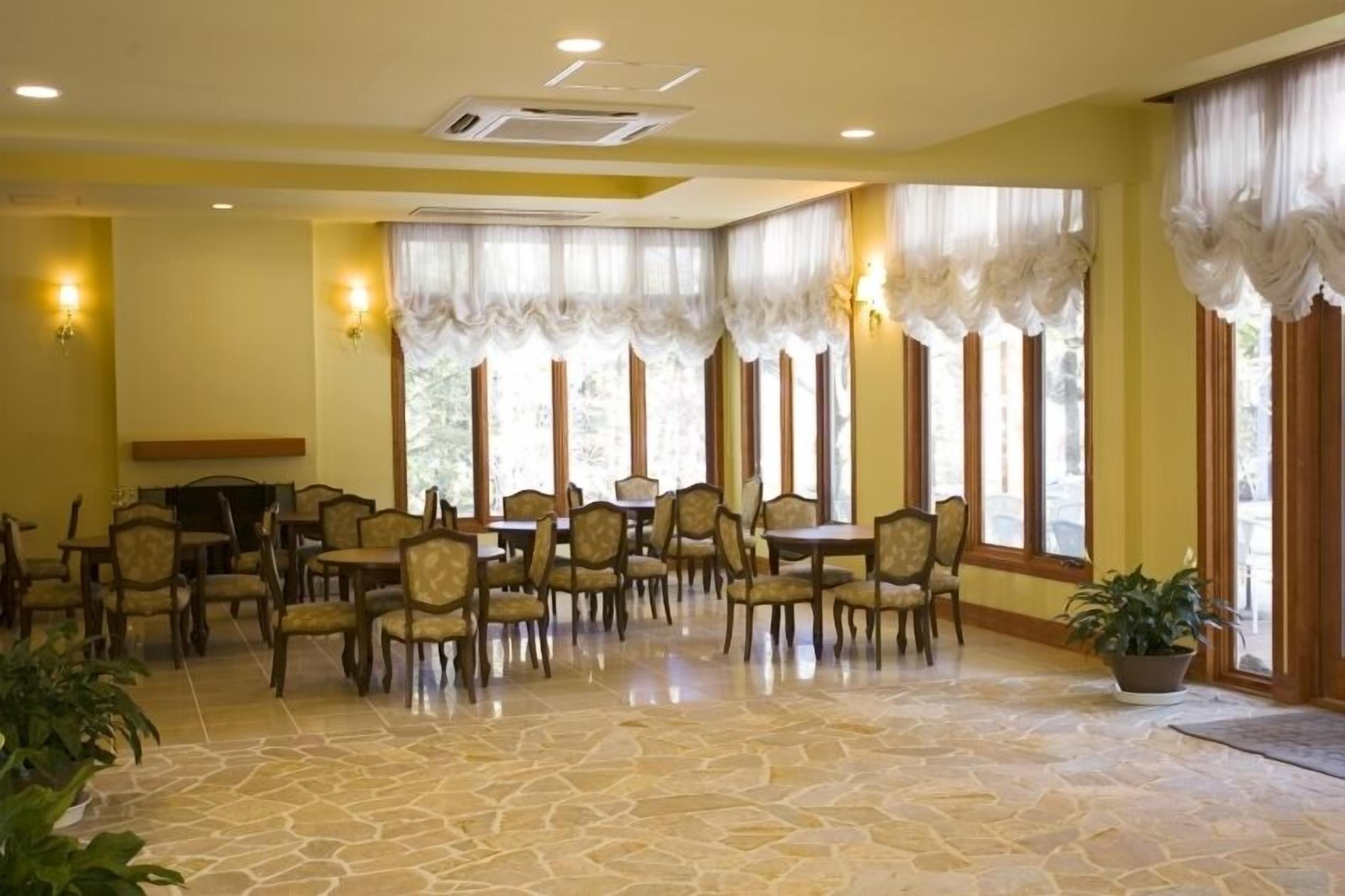 A dining area with stone flooring, multiple tables and chairs, and large windows with sheer curtains.