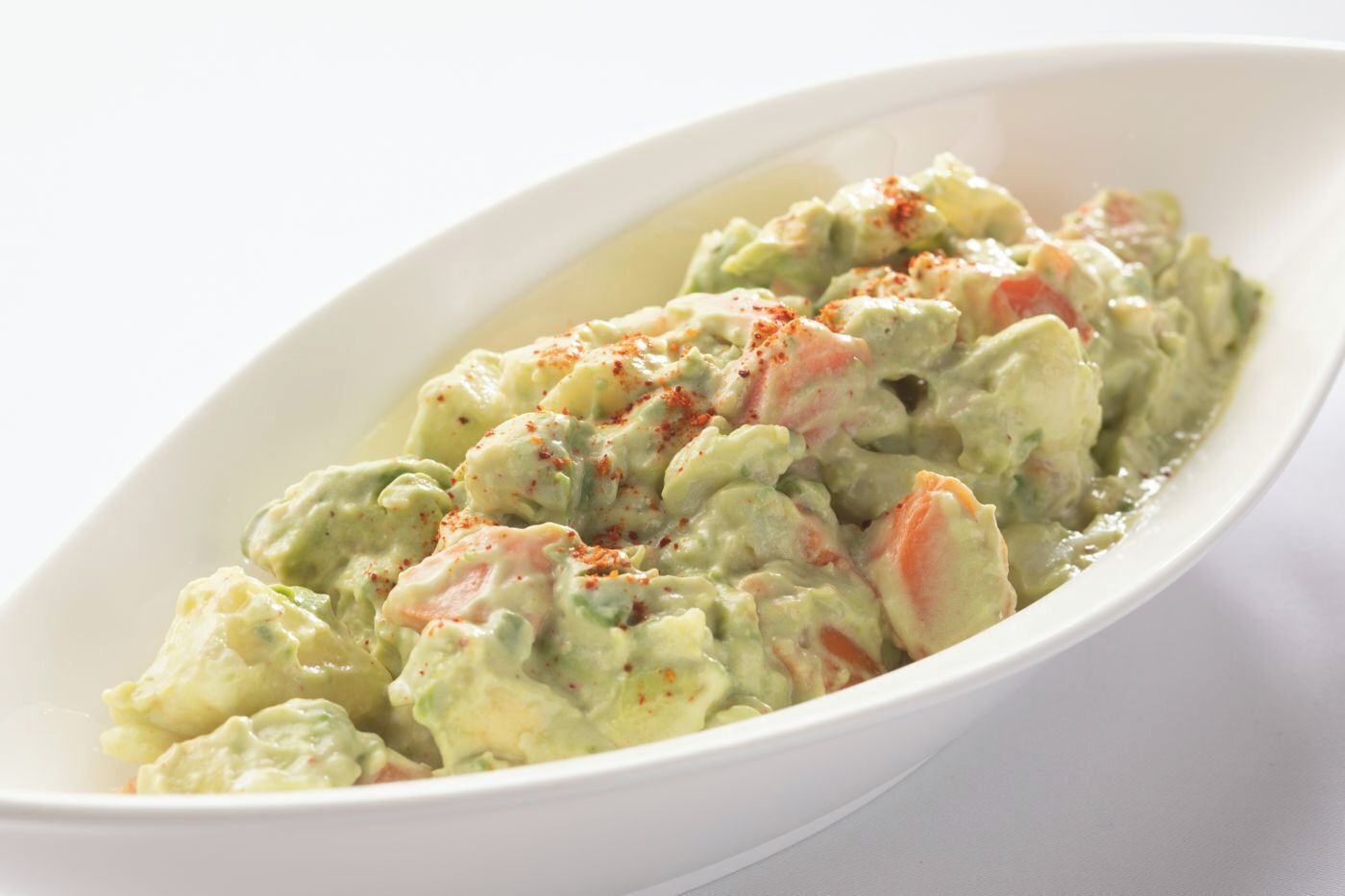 A white oval dish containing a creamy potato salad with visible chunks of potato, carrots, and a sprinkle of red seasoning.