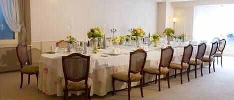 A long dining table set with chairs, candles, and floral centerpieces in a well-lit room with a large window.