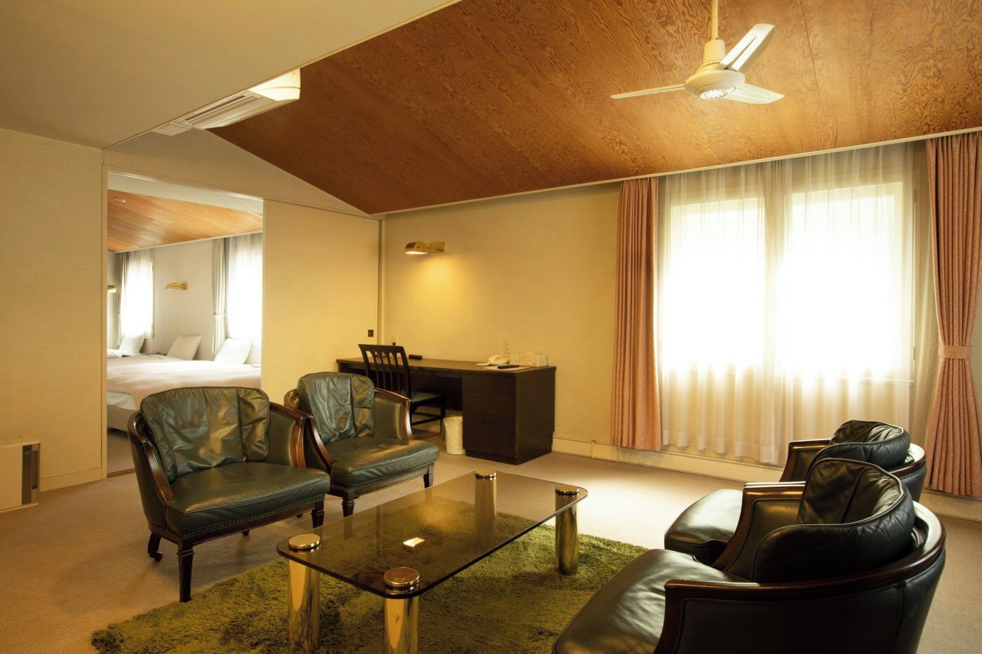 Habitación de hotel con cama, escritorio, dos sillones de cuero, una mesa de centro de vidrio y un ventilador de techo.