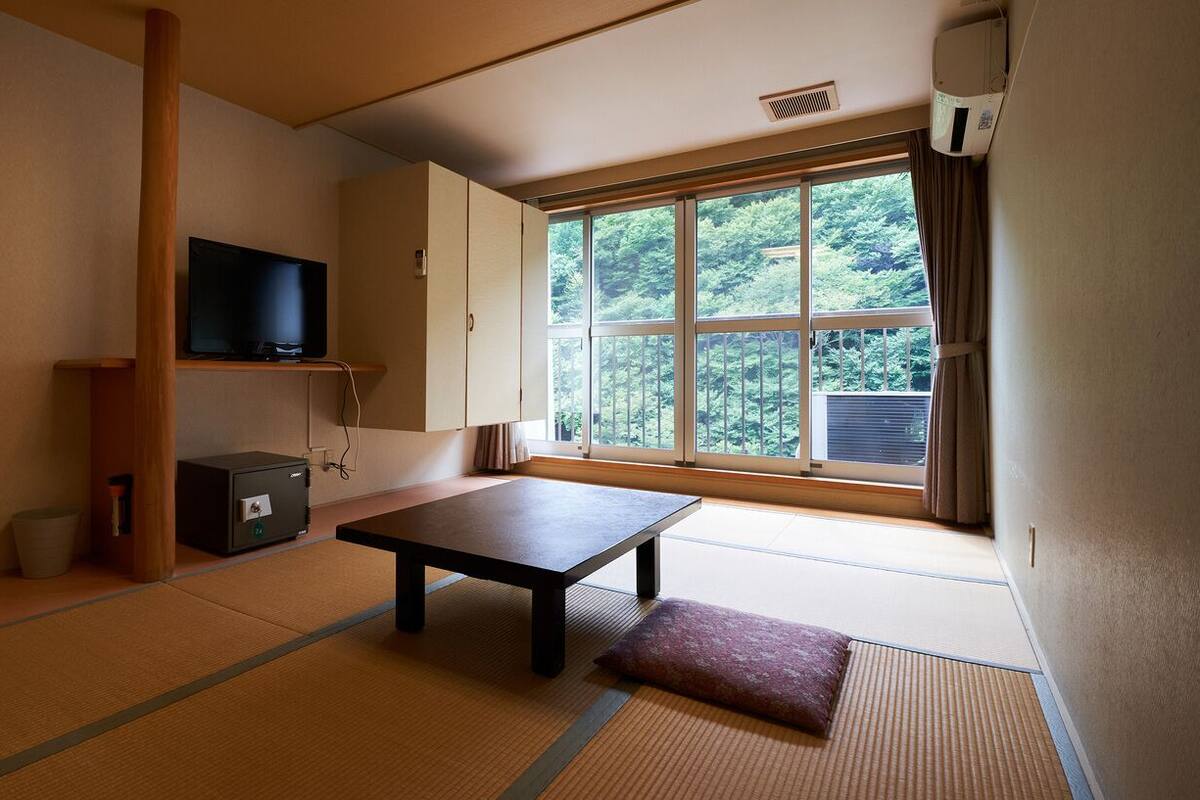 A traditional Japanese room with tatami flooring, a television, a cabinet, and a window with a view of trees.