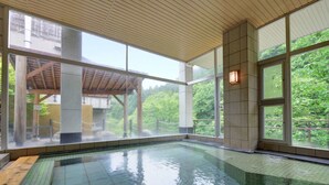 An indoor swimming pool with clear glass walls, wooden ceiling, and a view of greenery outside.