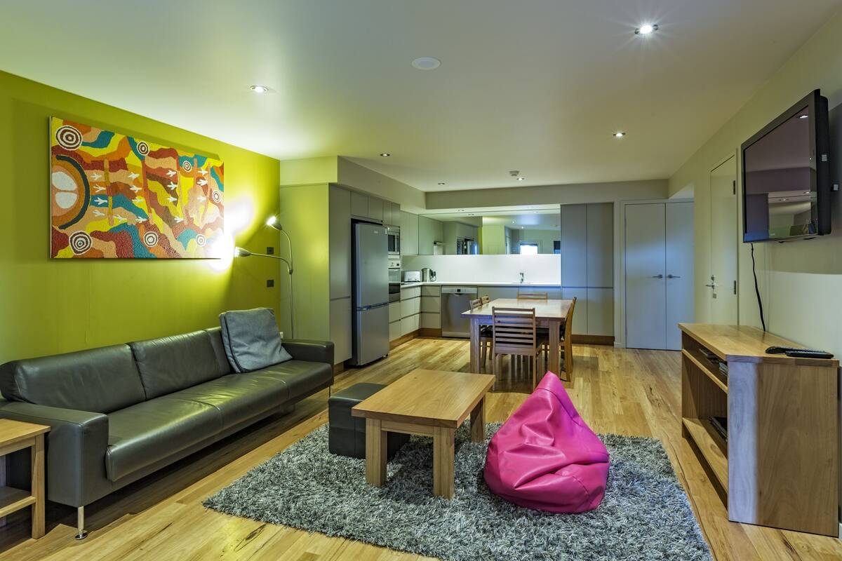 A modern living room with a sofa, coffee table, and bean bag. A kitchen is visible in the background with a dining area.
