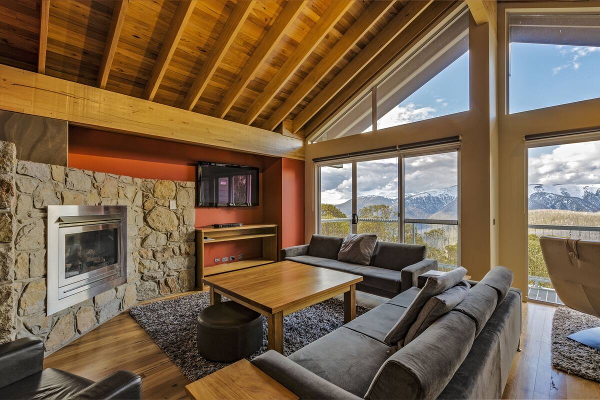 A modern living room with a stone fireplace, wooden ceiling beams, a stone wall, a large window with a mountain view, and a gray sofa.