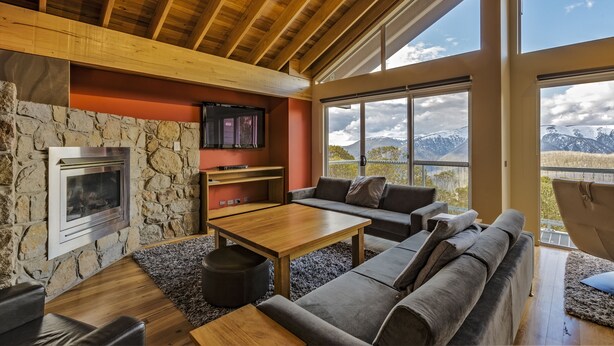 A modern living room with a stone fireplace, wooden ceiling beams, a stone wall, a large window with a mountain view, and a gray sofa.