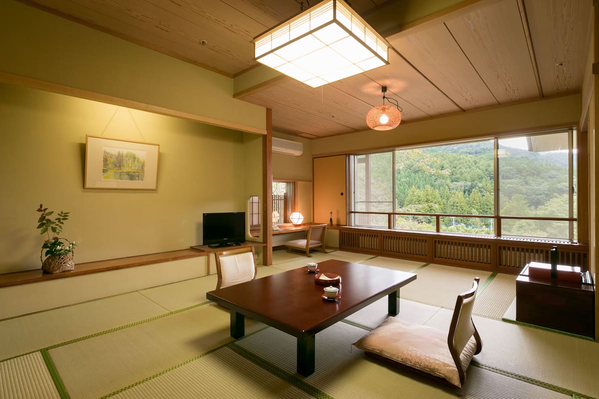 Una habitación japonesa tradicional con suelo de tatami, una mesa baja con cojines, un televisor y un amplio ventanal con vistas a una zona boscosa.