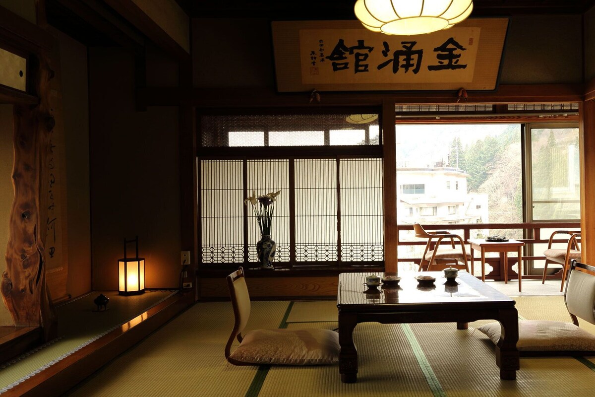 A traditional Japanese room with tatami flooring, a low table, chairs, a hanging lantern, and a view of the outside.